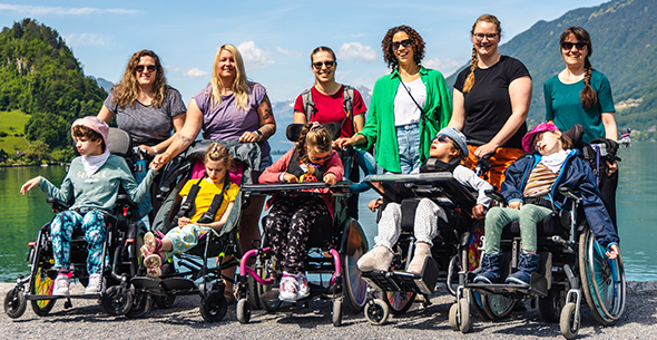 Fünf Schülerinnen, allesamt im Rollstuhl und ihre Lehr- und Betreuungspersonen posieren vor der Bergkulisse im Berner Oberland. Im Hintergrund sehen wir den grünen Brienzersee. 